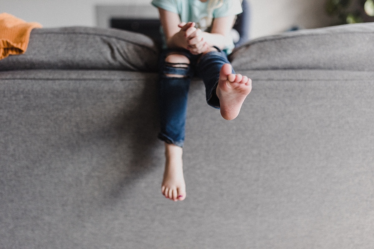 Little, swaying feet of a young girl in blue jeans sitting on the back of a couch