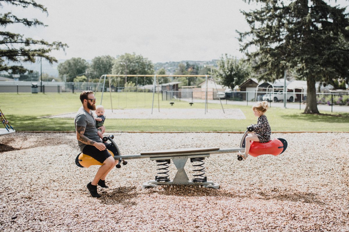 A father plays on the teeter totter with his kids at a park in Kamloops