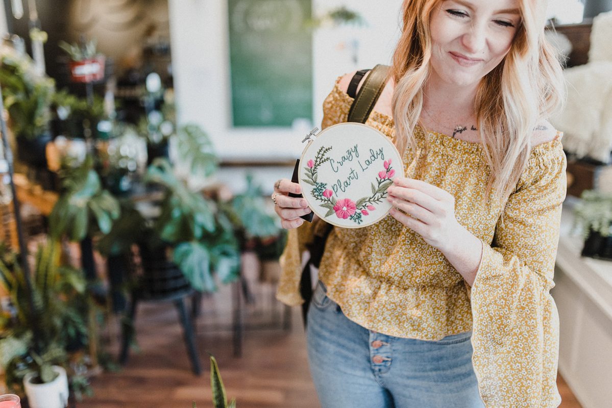 Skyleigh McCallum holding up an embroidered wall decor that reads "crazy plant lady" in local shop Far & Wide.