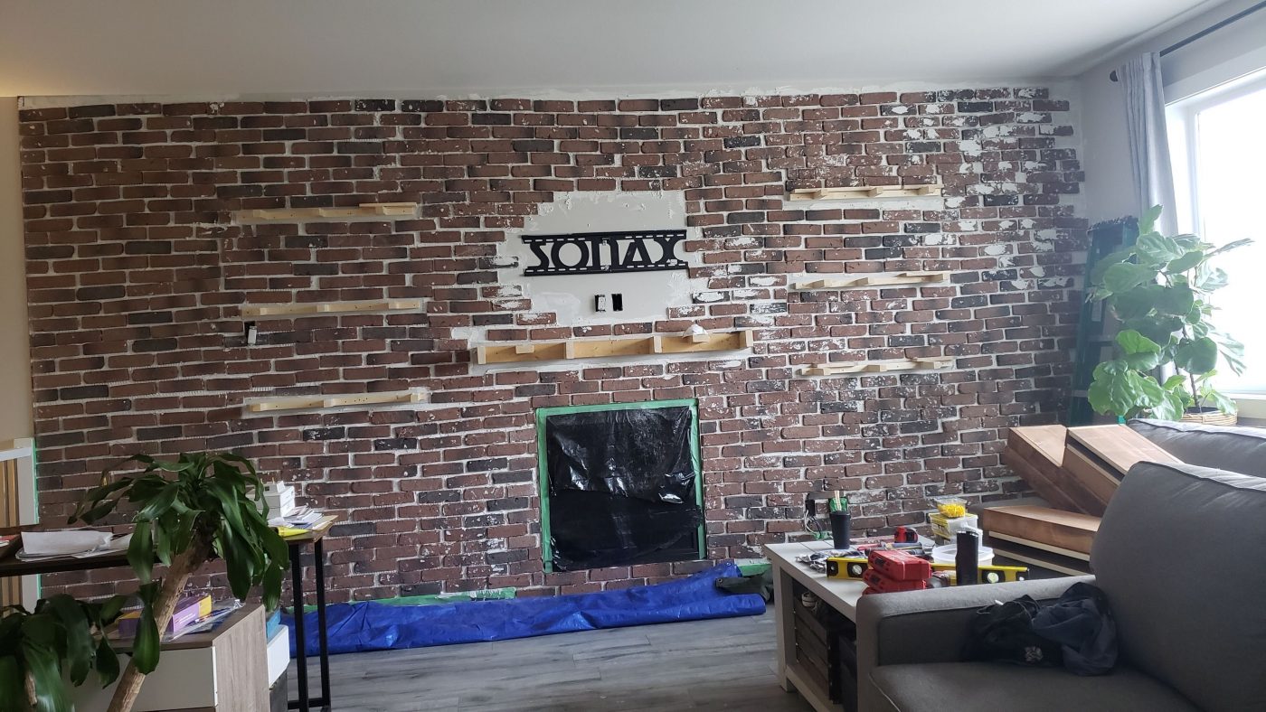 A do-it-yourself home renovation project in progress; faux brick tiles from Rona being placed on a plain wall with floating shelves and a fireplace.