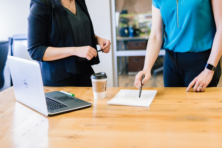 Two women stand in an office in front of a table with a laptop, coffee and notebook discussing opening an RRSP account to improve a credit score.