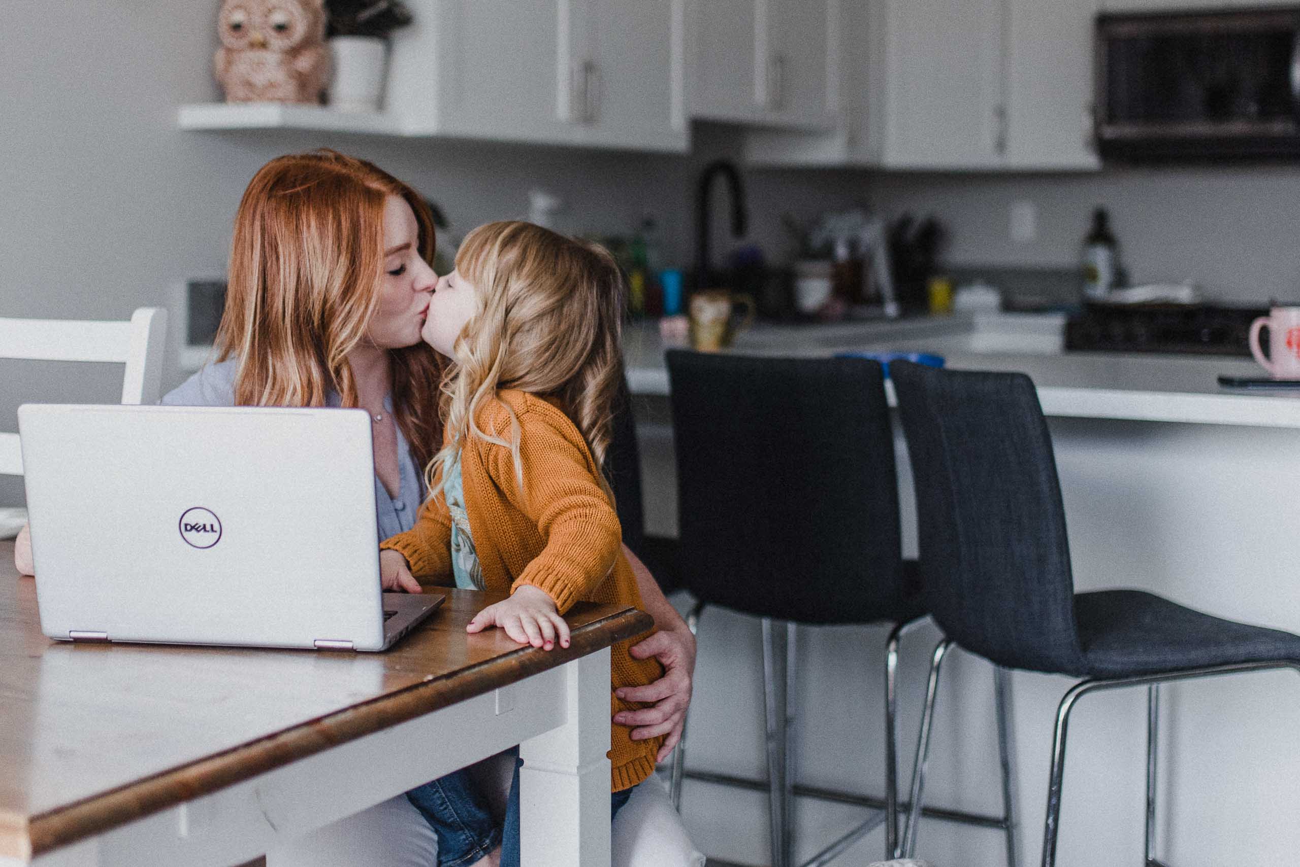Skyleigh McCallum, a Kamloops Realtor sits at the table in her kitchen in front of her laptop and gives her little daughter a smooch while their dog sits at her feet